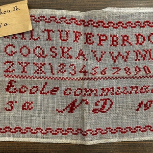 以下が含まれることがあります： 赤い刺繍でアルファベット、数字、そして「Ecole comme-ci-le」と「Jr D 1825」の文字が施された白いリネン製のサンプラーです。