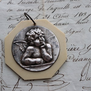 May include: A white octagonal pendant with a silver cherub design. The cherub is depicted with wings and is holding its chin in thought. The pendant has a loop at the top for hanging.