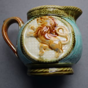 以下が含まれることがあります： 茶色と緑の釉薬をかけた小さな陶器のピッチャー。ピッチャーは、円形のフレームの中にライオンの浮き彫りの詳細なデザインが施されています。ピッチャーはハンドルと丸みを帯びたボディを持っています。