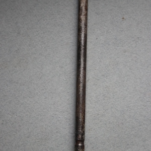 May include: A close-up of a metal sharpening steel with a wooden handle. The steel is silver and has a pointed tip. The handle is brown and has a silver band with a decorative pattern.