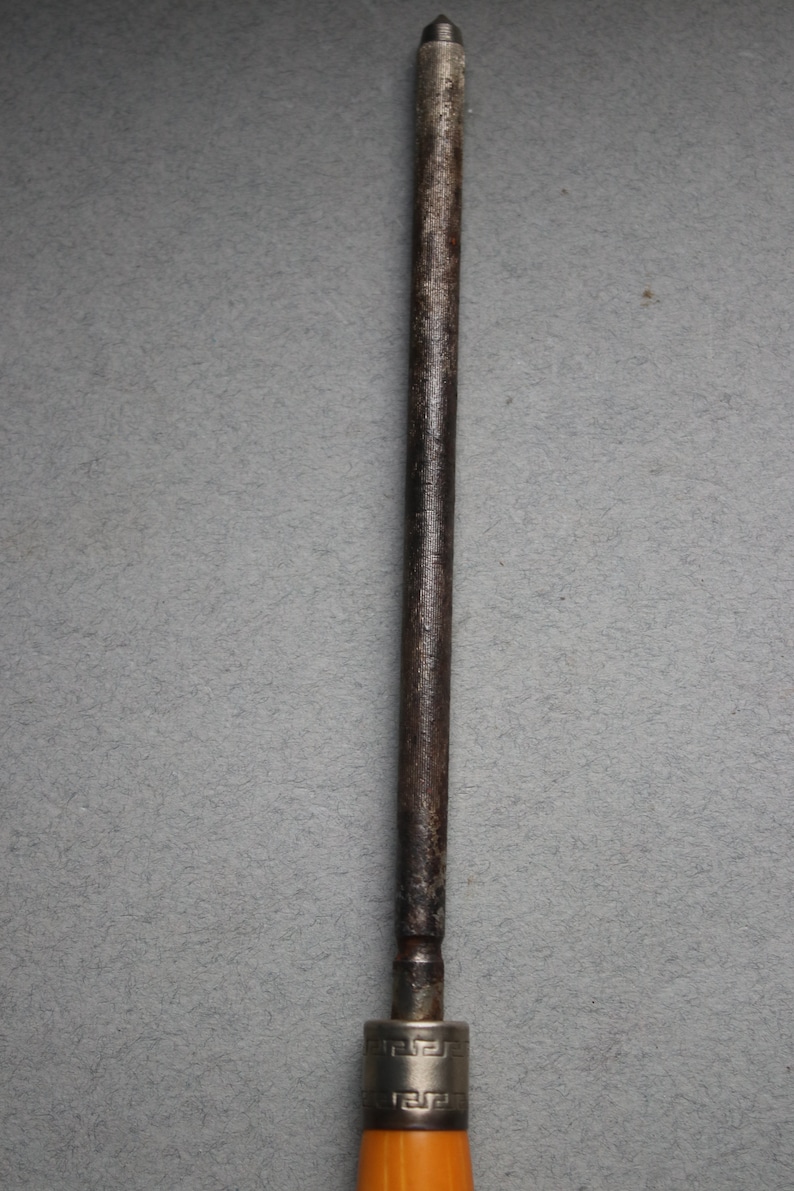May include: A close-up of a metal sharpening steel with a wooden handle. The steel is silver and has a pointed tip. The handle is brown and has a silver band with a decorative pattern.