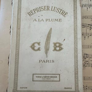 May include: A rectangular, antique box with the text "REPRISER LUSTRE A LA PLUME" and "PARIS" in gold lettering. The box features a feather illustration and the initials "C B". The box is on top of sheet music.