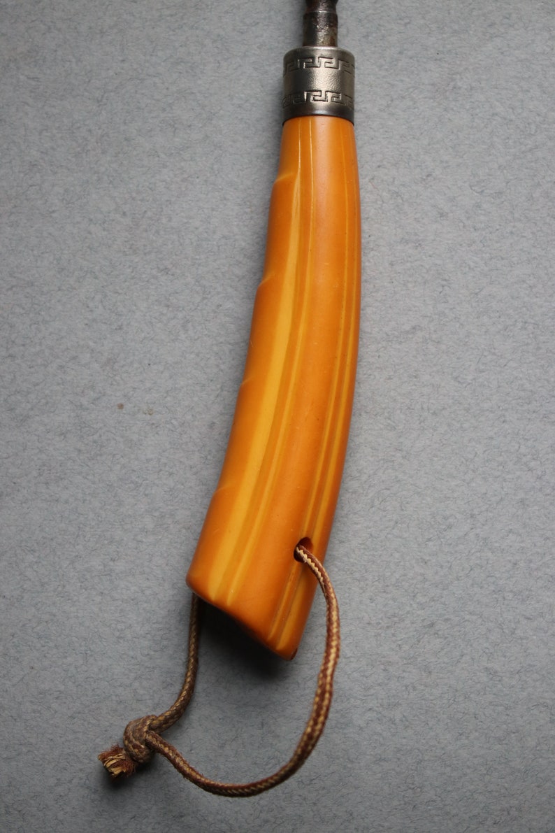 May include: A yellow and orange striped handle with a silver band and a Greek key design. The handle is attached to a brown cord.