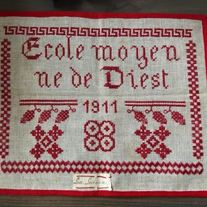 以下が含まれることがあります： 白いリネン生地に赤い刺繍が施されたサンプラー。テキストは「Ecole moyen nee de Diest 1911」です。