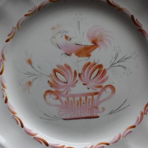 Pode incluir: Prato de cerâmica branco com um desenho de galo rosa e castanho. O galo está empoleirado em um vaso de flores rosa e castanho sobre um fundo branco. O prato tem uma borda ondulada e uma borda vermelha.