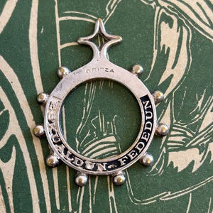 Peut inclure: Un pendentif circulaire argenté avec les inscriptions "ORITZA" et "FEDEDUN" gravées. Le pendentif est orné d'une étoile en haut et de petites décorations rondes sur le bord extérieur. Le fond est une surface à motifs verts et blancs.