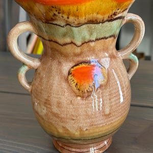 May include: A ceramic vase with two handles, featuring a color palette of brown, orange, yellow, and green. The vase has a bulbous body and a flared rim. The design includes a textured, layered effect, adding visual interest to the decorative piece.