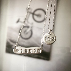 Puede incluir: Dos collares de plata con un colgante de plata. Un colgante es un rectángulo largo y delgado con los números "10516" grabados en él. El otro colgante es un disco redondo con los números "10:2" grabados en él.