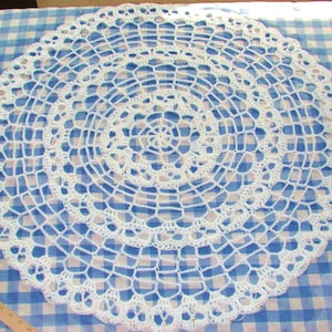 Puede incluir: Doily de ganchillo blanco con un diseño circular. El doily está colocado sobre una tela a cuadros azul y blanca.