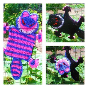 May include: Three crocheted cat figures hanging on a fence. The first cat is pink and blue with a wide grin. The second cat is black with a white grin. The third cat is black with a pink and blue grin.