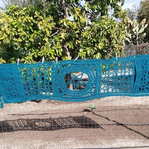 May include: A teal crocheted tapestry with a five-pointed star design in the center. The tapestry is hanging on a fence.