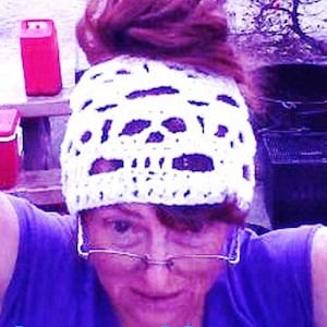 May include: A woman wearing a white crocheted hat with a spiderweb design. The hat is pulled up into a bun on top of her head. She is wearing a purple shirt and has glasses on.