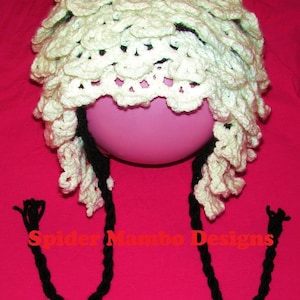 May include: White crocheted hat with a ruffled edge and two long black crocheted braids. The hat is displayed on a pink surface. The text "Spider Mambo Designs" is visible in the image.