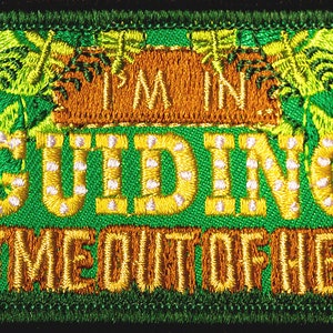 May include: Embroidered patch with a green background and the words "I'M IN GUIDING GET ME OUT OF HERE!" in yellow and orange. A blue and white trefoil emblem is in the upper left corner. The patch has a black border.