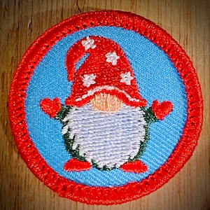May include: An embroidered gnome with a red hat and white stars on a blue background. The gnome is wearing red mittens and has a white beard. The gnome is surrounded by a red embroidered border.