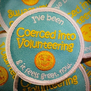 May include: A blue and white embroidered patch with the text "I've been coerced into volunteering & it feels great, now." and a yellow smiley face.