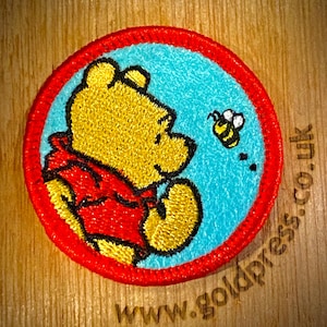 Op de afbeelding: Ronde geborduurde patch met een gele beer in een rood shirt, tegen een blauwe achtergrond en een rode rand. Er is ook een geel-zwarte bij aanwezig. De patch bevindt zich op een houten oppervlak met de tekst "www.goldpress.co.uk".
