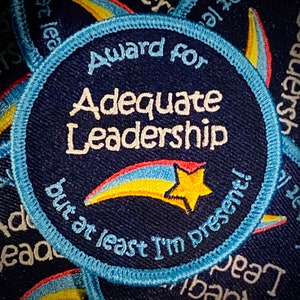Könnte beinhalten: Blau-weißer gestickter Aufnäher mit einem gelben Stern und einem Regenbogen. Der Text auf dem Aufnäher lautet "Award for Adequate Leadership but at least I'm present!"