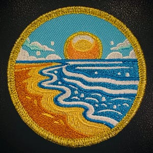 May include: A circular embroidered patch depicting a sunset over a beach scene. The sun is orange and yellow, the sky is blue, the beach is yellow and orange, and the water is blue with white waves.