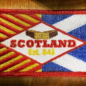 May include: Red, white, and blue embroidered patch with a diagonal white stripe and a yellow and red striped pattern. The patch features a diamond shape with the text "SCOTLAND Est. 843" and a brown and yellow waffle-like image.