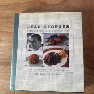 May include: A cookbook titled "JEAN-GEORGES Cooking at Home with a Four-Star Chef" by Jean-Georges Vongerichten and Mark Bittman. The cover features a portrait of the chef and images of prepared dishes. The book has a light blue spine.