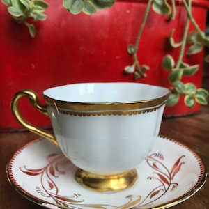May include: A white teacup and saucer with gold trim and a red and gold floral design. The teacup has a scalloped edge and the saucer has a scalloped rim.
