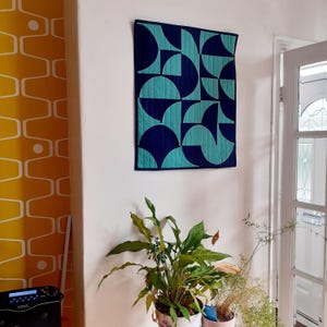 May include: A geometric wall hanging featuring a pattern of teal and navy blue semi-circles. The quilt is rectangular with a navy blue border and is mounted on a white wall. The design creates a modern, abstract aesthetic.