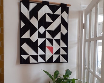May include: A geometric wall hanging with a black and white pattern, featuring a single red triangle. The textile is mounted on a wooden dowel with black fabric loops. The design is modern and abstract.