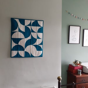 Op de afbeelding: Een gewatteerde wandhanger met een geometrisch ontwerp van turquoise en witte halve cirkels. De quilt is rechthoekig en omlijst met een turquoise rand. Het hangt aan een grijze muur en voegt een moderne touch toe aan de kamerdecoratie.