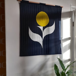 May include: A navy blue wall hanging with a yellow sun-like shape and white floral design. The fabric is quilted with vertical lines and is mounted on a wooden rod. The wall hanging is displayed on a white wall.