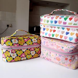 Könnte beinhalten: Drei Kosmetiktaschen aus Stoff mit Reißverschlüssen und Tragegriffen. Die Taschen weisen farbenfrohe Blumen- und Herzmuster auf weißem Hintergrund auf. Die Taschen sind übereinander gestapelt, die größte Tasche unten.