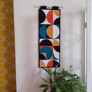 May include: A geometric wall hanging featuring a repeating pattern of orange, teal, yellow, and white semi-circles against a black background. The artwork is suspended from a wooden dowel. A potted plant is visible below.