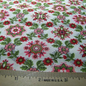 May include: Close-up of a white fabric with a repeating floral pattern. The design features pink, red, and green flowers and leaves. A wooden ruler with inch markings is in the foreground, with the text "MADE IN U.S.A." visible.