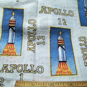 May include: White fabric with a repeating pattern of blue and white rockets with yellow flames. The rockets are labeled "Apollo" with a number. The fabric has the text "Apollo 1" printed on it.