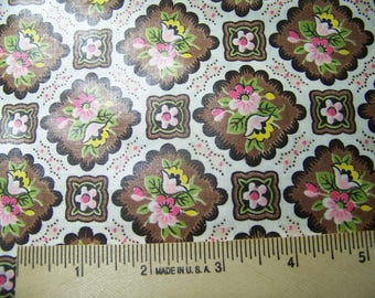 May include: Close-up of a fabric with a repeating floral pattern. The design features brown, scalloped squares with pink and yellow flowers, green leaves, and smaller square floral accents. The background is white with pink dots. A ruler is at the bottom.