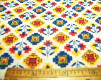 May include: White fabric with a repeating pattern of blue, red, and yellow geometric shapes. The design includes square and diamond shapes, with floral and star-like elements. A ruler is at the bottom.