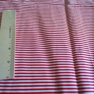 May include: A piece of fabric featuring a pattern of thin, horizontal red and white stripes. A wooden ruler is placed vertically along the left side, showing a measurement scale that extends to 6 inches in length for scale and reference.