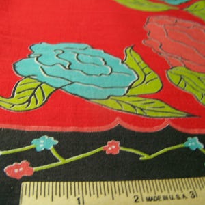 May include: Close-up of a red fabric with a floral pattern. Large turquoise flowers with green leaves are visible, along with a border of small flowers on a black background. A ruler is at the bottom, marked in inches.