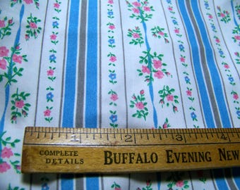 May include: White fabric with blue and gray stripes and a repeating pattern of pink and blue flowers. A wooden ruler with the text "COMPLETE DETAILS" and "BUFFALO EVENING NEWS" is placed on top of the fabric.