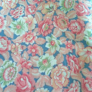 May include: Close-up of a floral fabric with a repeating pattern of pink, peach, and green flowers on a blue background. The flowers are in various stages of bloom, with detailed petals and leaves.