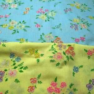 May include: Two pieces of floral fabric. One is light blue with clusters of pink, yellow, and purple flowers. The other is yellow with similar floral patterns. Green leaves accent the flowers.