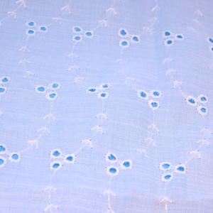 May include: Close-up of a light blue fabric with a delicate eyelet pattern. The design features small, three-petal flower shapes with blue centers, connected by white, arrow-like lines. The fabric has a soft, textured appearance.