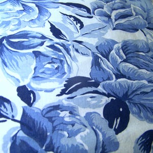 May include: Close-up of a fabric with a floral pattern. The design features large, stylized roses in shades of blue against a white background. The roses have multiple layers of petals and are surrounded by dark blue leaves. The overall effect is a classic, elegant design.