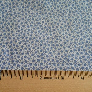 May include: Close-up of fabric with a repeating pattern of small blue flowers on a white background. A wooden ruler, marked in inches, is at the bottom, indicating the fabric's scale. The fabric is likely cotton.