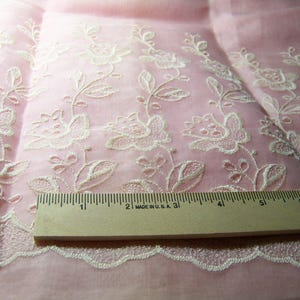 May include: Close-up of pink fabric with white floral embroidery. A wooden ruler, marked in inches, rests on the fabric. The embroidery features detailed flowers and leaves along the edges, with a delicate, scalloped lace trim. The fabric is slightly translucent.