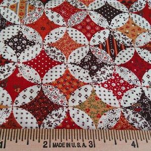 May include: Close-up of a fabric with a repeating pattern of interlocking circles and diamonds. The fabric features a mix of red, brown, white, and tan colors. The pattern includes floral and polka dot designs. A ruler is at the bottom.