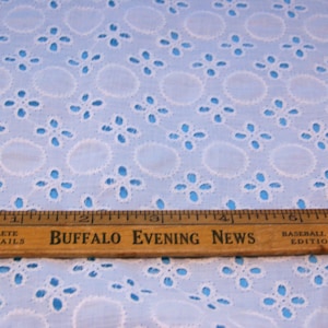 May include: A light blue eyelet fabric with a repeating pattern of white embroidered circles and floral designs. A wooden ruler with the text "Buffalo Evening News" and "Baseball Edition" is placed across the fabric.