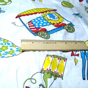 May include: White fabric with a colorful circus-themed print. The design includes a lion in a wagon, a blue giraffe, and other whimsical elements. A wooden ruler is placed on the fabric, measuring approximately 5 inches.