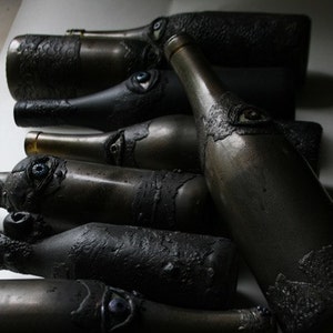 May include: A collection of dark gray glass bottles with textured surfaces and decorative eyes. The bottles are arranged in a row, with the largest bottle in the center.