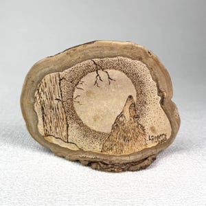 May include: A circular wood slice featuring a pyrography design of a wolf howling at a large moon. The artwork includes a tree silhouette on the left, stippled shading around the moon, and the signature L. Rogers 2006 in the bottom right corner.
